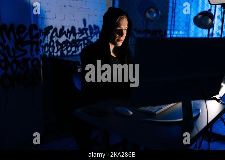 Medium shot portrait of hidden hacker man wearing sweatshirt with hood engaged in hacking into security systems in dark basement room with blue neon Stock Photo