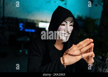 Close-up face of confident wanted hacker man wearing anonymous mask and hoodie holding beads in hands and looking away sitting at table in basement Stock Photo