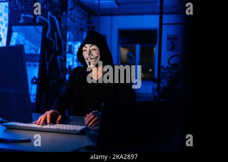 Portrait of unrecognizable dangerous hacker man in anonymous mask starting breaking computer system and typing password for hacking bank account. Stock Photo