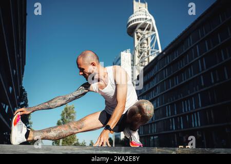 Young muscular tattooed man doing exercises, stretching early in the morning, bald bearded guy in sportswear stretching legs before jogging Stock Photo