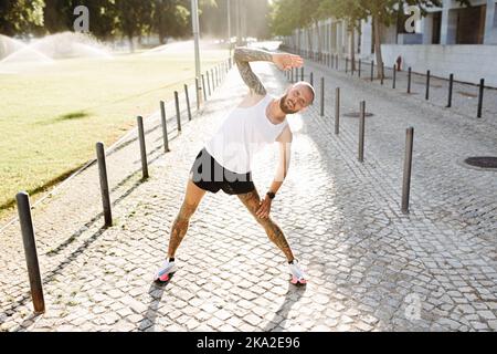 Athletic young tattooed man warming up by stretching before exercises outdoors, bearded fit guy in sportswear doing training exercises Stock Photo