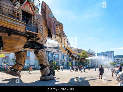 The Great Elephant giant puppet, part of the Machines of the Isle of Nantes, sprays water with its trunk on onlookers along of the shipyards building. Stock Photo