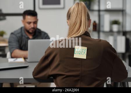 Back of young blond businesswoman with word loser written on sticky notepaper sitting by workplace in front of Hispanic male colleague Stock Photo