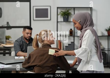 Young smiling Muslim woman in hijab putting notepaper with word fool on back of blond businesswoman sitting by workplace Stock Photo