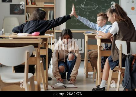 Two boys giving each other high five over African American classmate taking her copybook in aisle between rows of desks Stock Photo