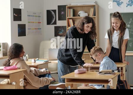 Group of youthful schoolchildren bullying junior schoolboy drooping his head while sitting by desk among elder pupils making fun of him Stock Photo