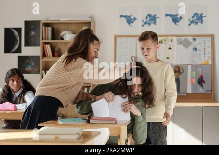 Cruel schoolgirl laughing at classmate and pulling his hair while another boy fighting with him in classroom against African American girl Stock Photo