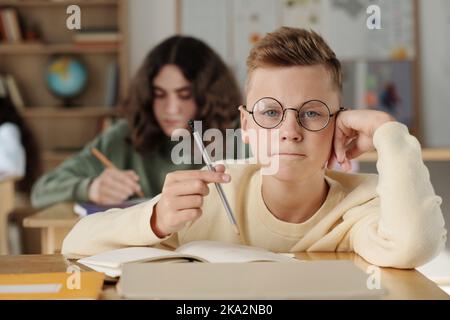 Cute clever secondary school learner in eyeglasses holding pencil over open copybook and looking at camera during individual work at lesson Stock Photo