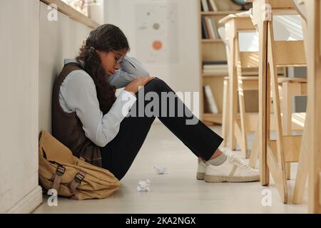 Side view of lonely and upset African American schoolgirl sitting on the floor of classroom while suffering from bullying of her classmates Stock Photo