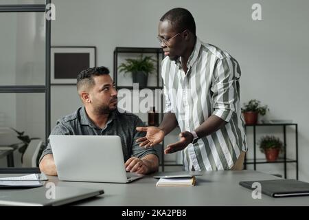 Young irritated African American employee looking at multi-ethnic or Hispanic man sitting by desk in front of laptop during conversation Stock Photo