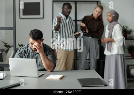 Middle aged Hispanic businessman sitting by workplace and networking against group of cruel intercultural colleagues bullying him Stock Photo