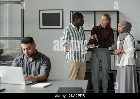Group of young multicultural colleagues talking about Hispanic male coworker networking by desk while standing behind him Stock Photo