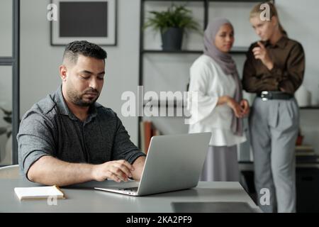 Young Hispanic businessman networking or preparing project in front of laptop against two intercultural female colleagues bullying him Stock Photo