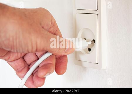 Close up of hand pulling cord out or in of modern white outlet on wall. Plug from an electrical appliance plugged into an electrical outlet on a white Stock Photo