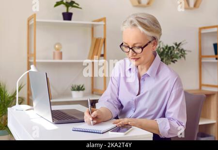 Senior businesswoman is making notes in notewbook sitting at desk with laptop and smartphone. Stock Photo