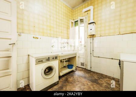 Dilapidated old dirty kitchen with dark floor, white tiles and yellow wallpaper on the walls, lonely cooker and gas heater, one piece sink and vintage Stock Photo