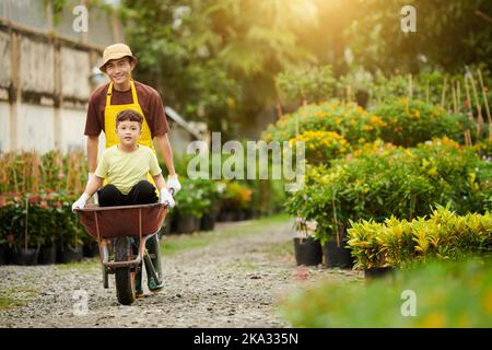 Little boy sitting inside wheelbarrow when father pushing it along path at plant nursery Stock Photo