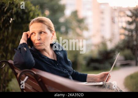 Mature business woman holding laptop talking on mobile phone in park Stock Photo