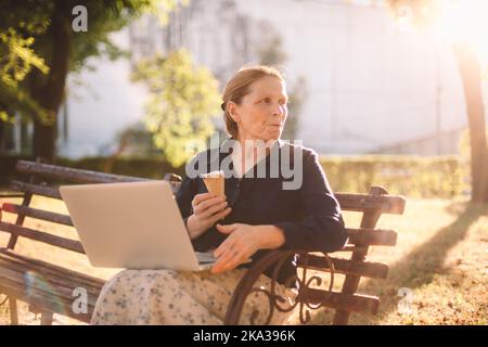 Mature business woman eating ice cream using laptop computer in park Stock Photo