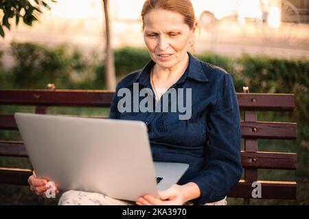 Mature woman using laptop computer having video call sitting on bench Stock Photo