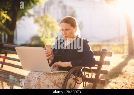 Mature business woman eating ice cream using laptop computer in park Stock Photo