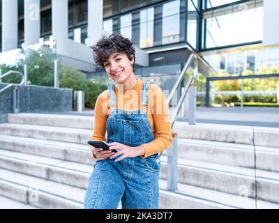 Smiling ethnic female sitting on railing on railing of porch browsing on smartphone near modern building on city street in sunlight Stock Photo