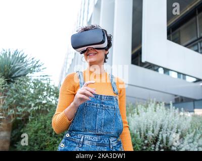Cheerful woman in VR goggles interacting with virtual reality while playing video game on sunny day Stock Photo