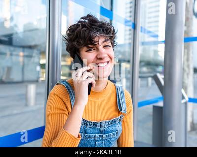 Smiling young female with short curly hair in denim overall standing near glass wall of urban building and making phone call at street Stock Photo