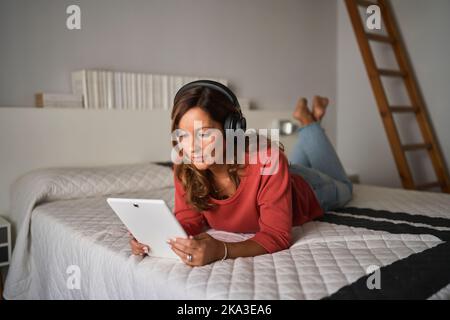 Positive middle aged Hispanic woman listening to music and browsing social media on tablet while lying on bed on weekend day at home Stock Photo