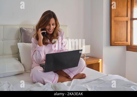 Middle aged Hispanic woman in pink pajama browsing data on laptop and speaking on cellphone while sitting cross legged on bed in sunlit bedroom in mor Stock Photo