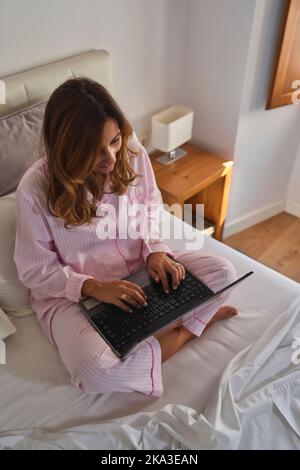From above middle aged Hispanic woman in pink pajama browsing data on laptop while sitting cross legged on bed in sunlit bedroom in morning at home Stock Photo