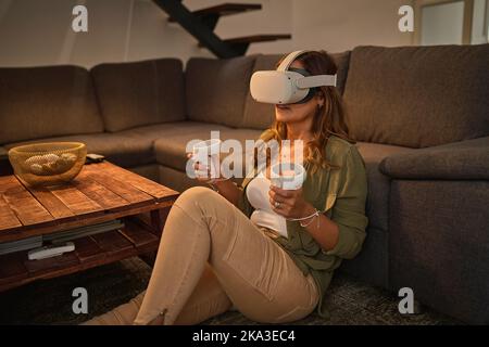 Mature female gamer in stylish clothes with controllers sitting on floor near sofa and playing VR videogame in weekend evening in living room at home Stock Photo