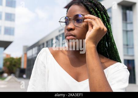 Young cool African American woman in white top with long braided hair wearing eyeglasses and looking away with confidence in town Stock Photo