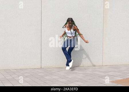 Full body of young stylish African American female with long braided hair in casual top and denim dancing on pavement near wall Stock Photo