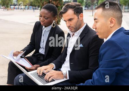 Group of multiracial coworkers in formal suits sitting on bench and working with laptop and documents while looking at screen Stock Photo
