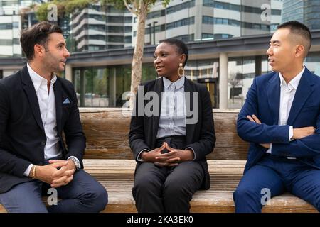 Positive multiracial coworkers in formal suits sitting on wooden bench near modern business center and chatting during break Stock Photo