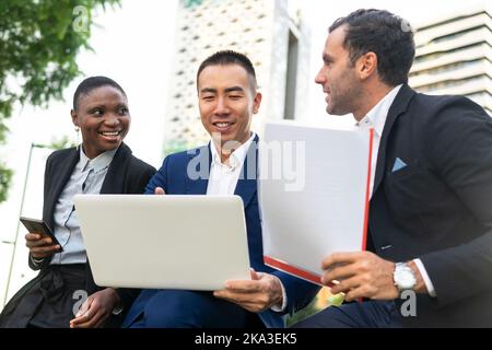 Group of smiling multiracial coworkers in formal suits sitting on bench and working with laptop and documents while looking at screen Stock Photo