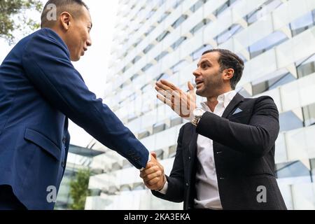 Low angle of positive multiracial businessmen in formal suits standing on street near modern multistory building and talking while shaking hands Stock Photo