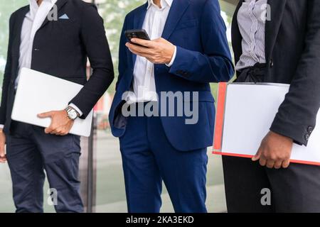 Cropped unrecognizable diverse coworkers in formal clothes standing near glass wall and looking at smartphone screen together while holding folder and Stock Photo