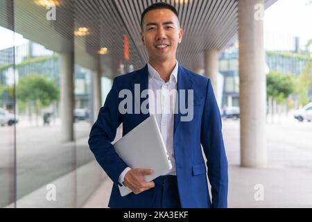 Positive Asian male entrepreneur in formal suit standing under entrance canopy near glass wall of modern building and holding laptop while looking at Stock Photo