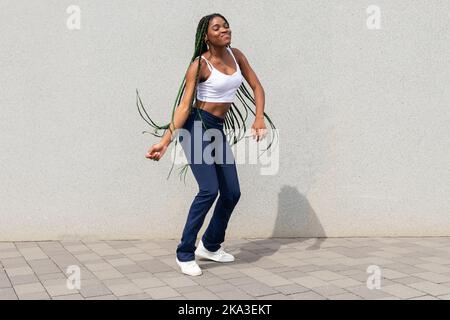Full body of young stylish African American female with long braided hair in casual top and denim dancing on pavement near wall Stock Photo