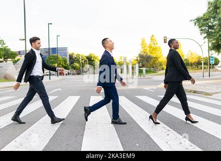 Side view full body of cheerful diverse coworkers in formal clothes walking along pedestrian crossing and chatting on street Stock Photo