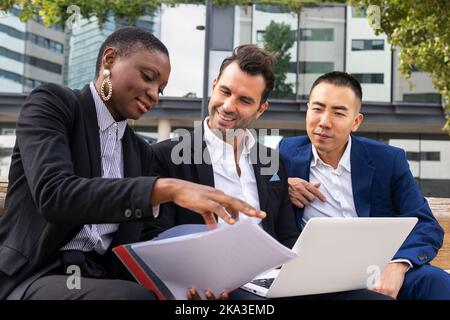 Group of multiracial coworkers in formal suits sitting on bench and working with laptop and documents while looking at screen Stock Photo