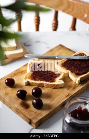 High angle of appetizing crispy toasts with cherry jam served on wooden chopping board near fresh ripe cherries on table Stock Photo