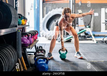 Full body of young fit female athlete in sportswear lifting kettlebell while training in gym Stock Photo