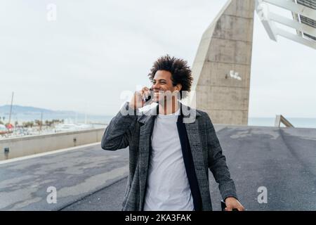 African American male traveler walking with suitcase talking on smartphone during vacation in city Stock Photo