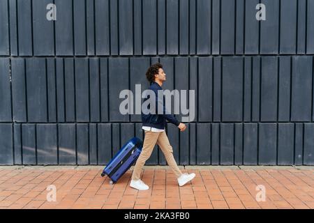 Full body African American male tourist with curly hair pulling suitcase and walking on pavement against gray wall of modern building Stock Photo