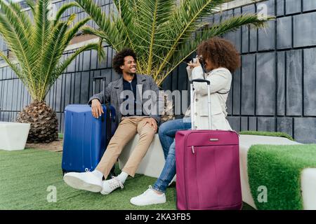 Full body African American female traveler in outerwear with curly hair smiling and shooting cheerful boyfriend near suitcase while sitting on bench n Stock Photo