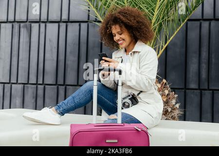 Side view of young African American female in casual clothes sitting on bench messaging on mobile phone near luggage Stock Photo
