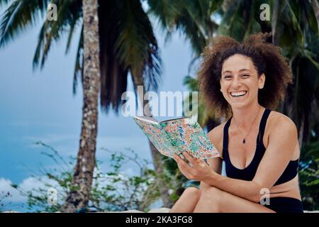 Smiling focused Hispanic female in black top sitting with journal on tropical beach with palm trees and looking at camera Stock Photo